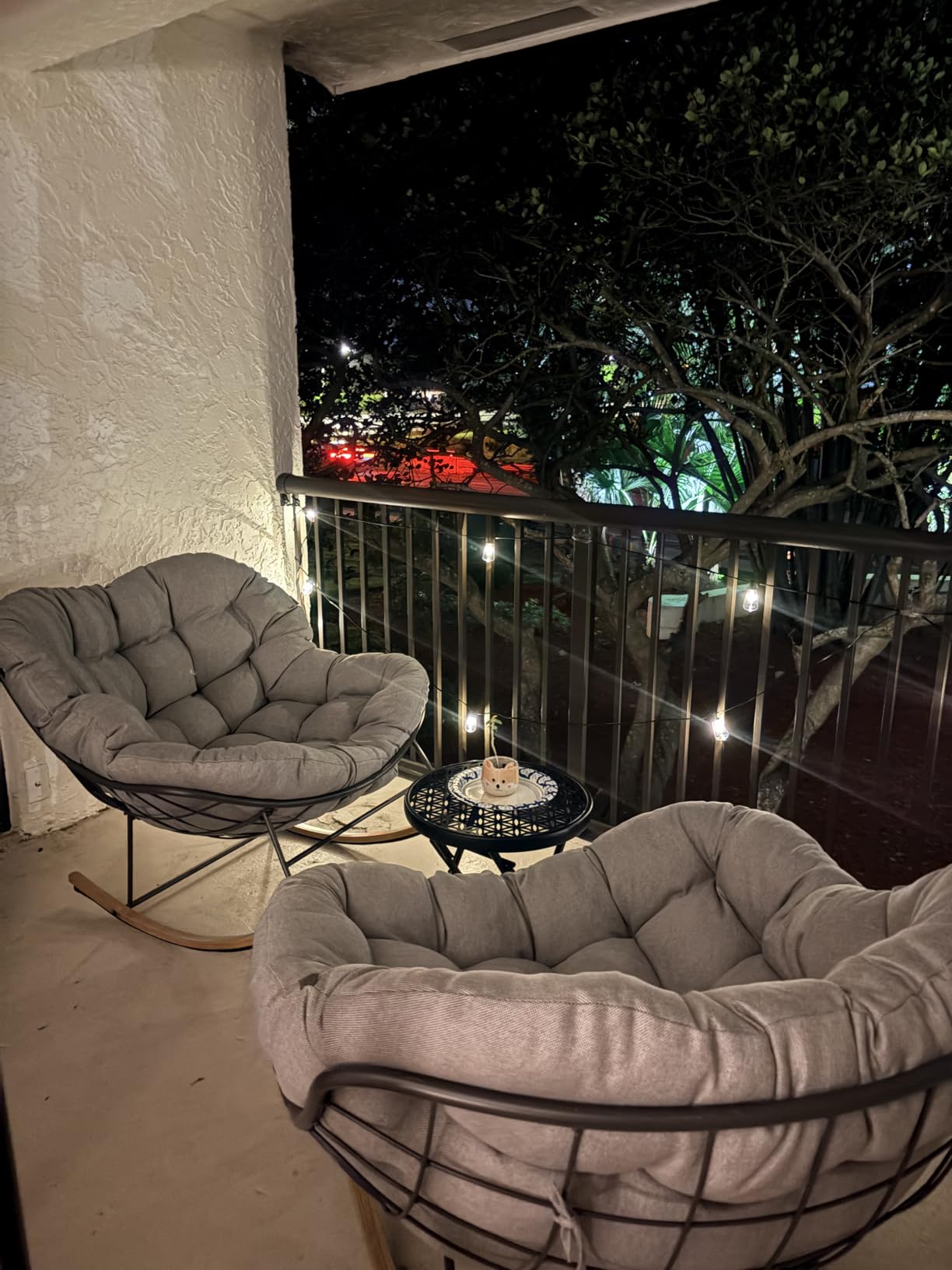 Two oversized cushioned rocking chairs set around a small round lattice-top table with a ceramic planter on an apartment balcony at night, string lights along the railing