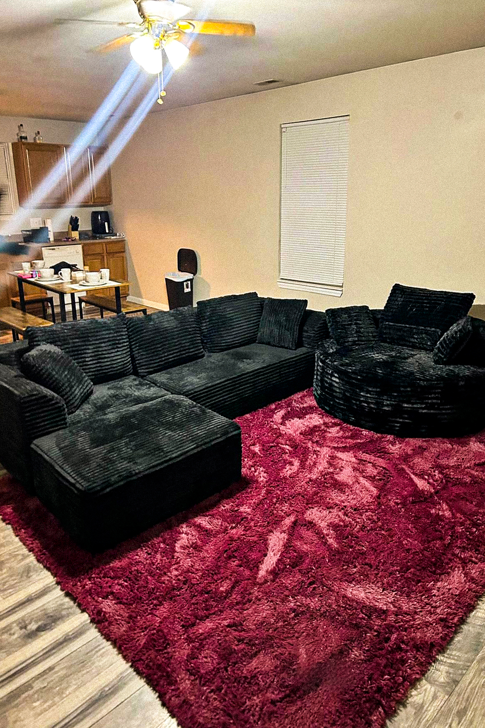 Black corduroy L-shaped sectional with a chaise and a round black corduroy swivel chair on a deep maroon shag rug, in an open plan living room with beige walls, a ceiling fan, and a kitchen with a small dining table behind