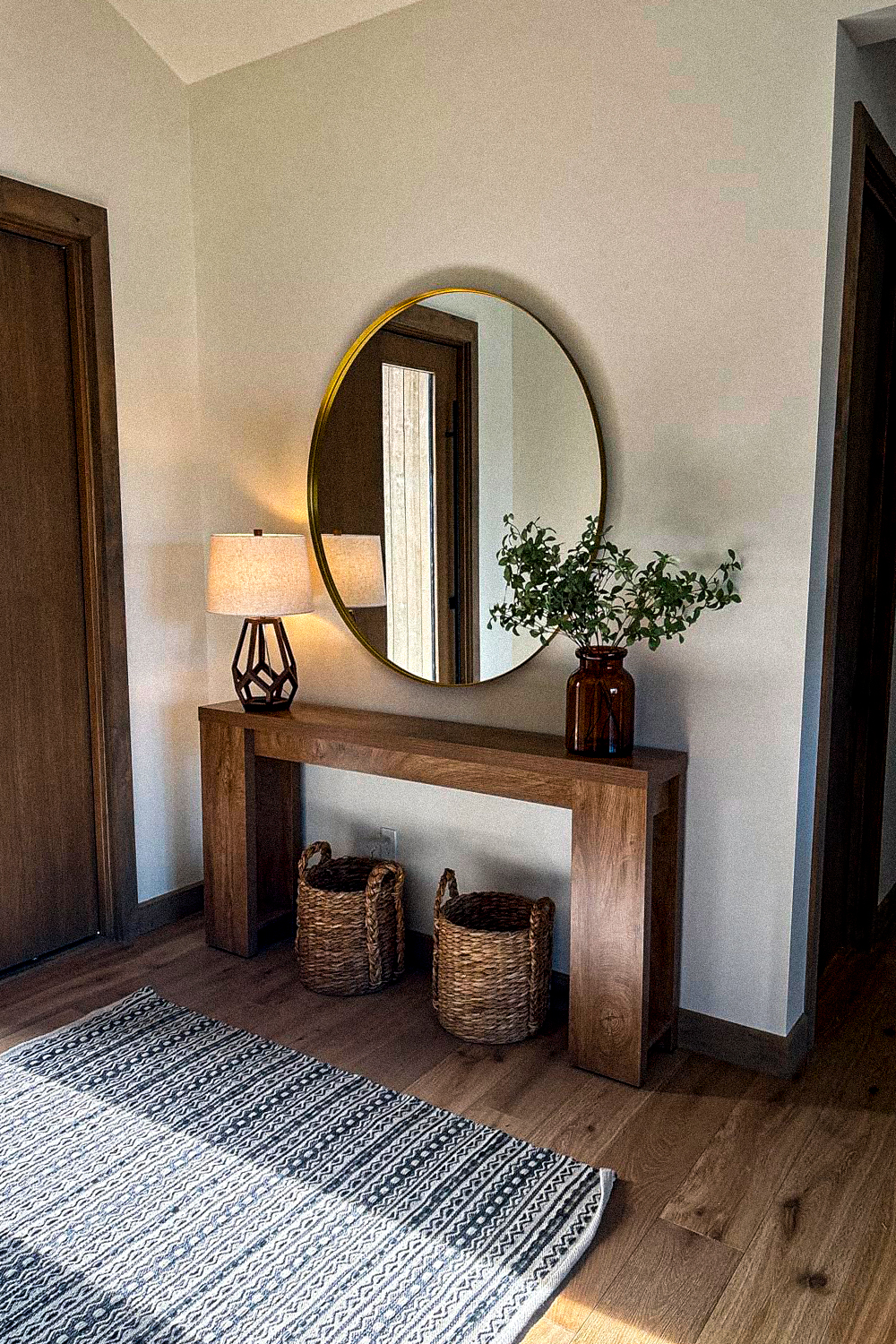 Chunky open wood console table with thick slab legs under a round thin brass-frame mirror, holding a lamp with a dark geometric base and cream shade and an amber glass vase with greenery, two woven seagrass baskets with handles underneath, and a grey and white striped runner rug on a wood floor beside a dark wood door