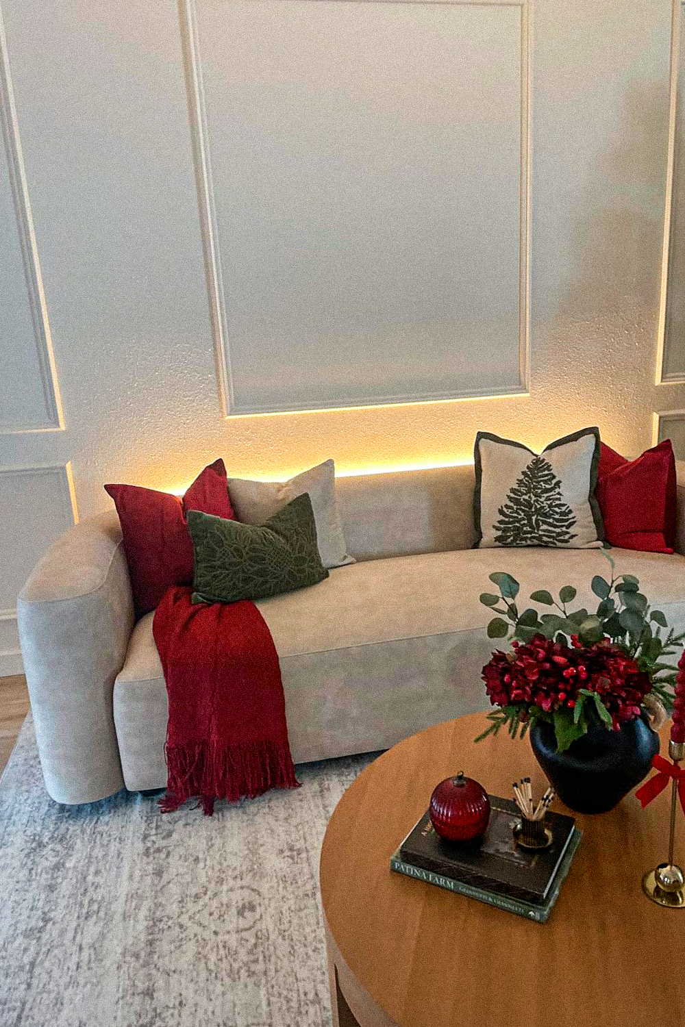 Curved cream sofa with red, forest green and pine tree embroidered throw pillows and a red fringed throw, against a paneled wall with warm LED backlight, beside a round wood coffee table with a black vase of red flowers and a stack of books, on a grey vintage-style rug