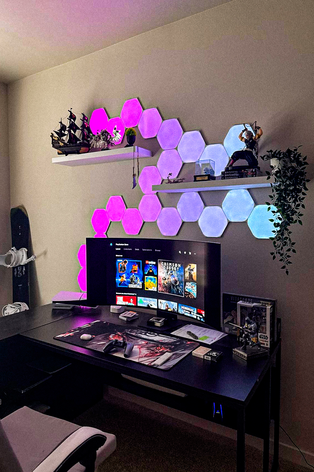 Hexagon LED light panels glowing pink, purple and blue on a wall above a black gaming desk with a curved monitor, an extended desk mat and a controller, white floating shelves with a model ship and figurines, and a trailing plant in the corner