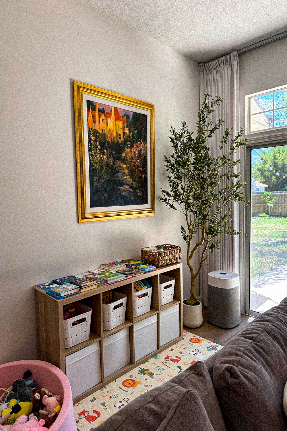 Artificial olive tree in a white pot beside a sliding glass door, next to an eight-cube storage shelf with white bins and a framed landscape print