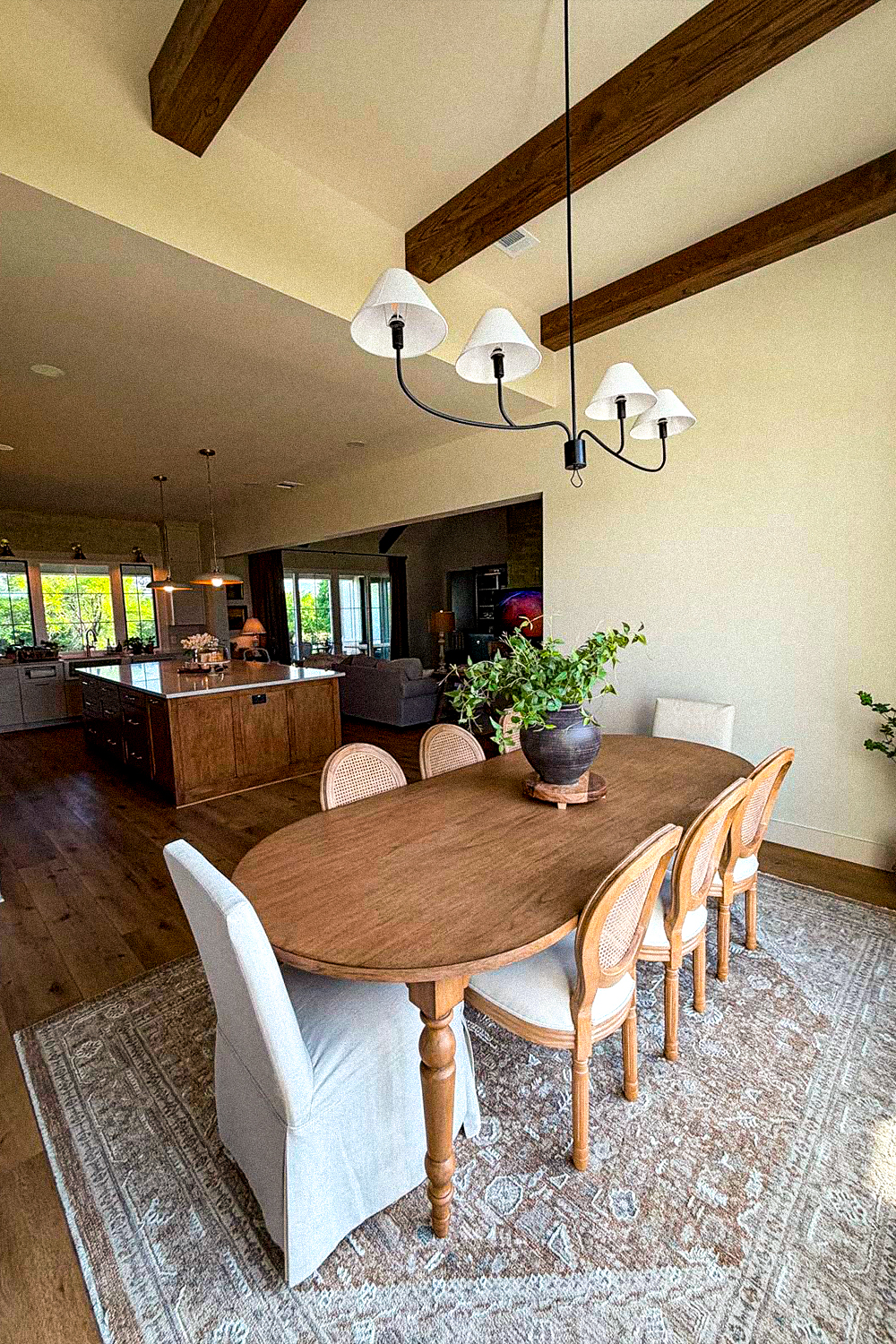 Long oval wood dining table on turned legs with french cane back chairs along the sides and two light grey slipcovered chairs at the ends, on a faded vintage-style rug, under a black chandelier with white shades and dark exposed beams on a vaulted ceiling, with a kitchen island beyond