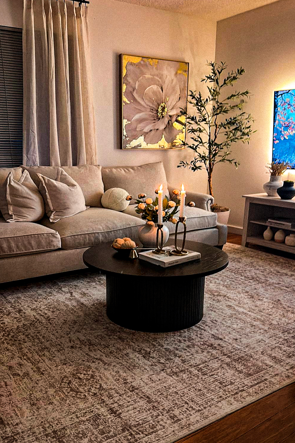 Round black fluted drum coffee table with two lit taper candles in brass holders, a small vase of dried flowers and a book, on a faded mauve and grey distressed rug, in front of a cream sectional with pillows, under a large floral canvas print, beside a faux olive tree and a low tv console