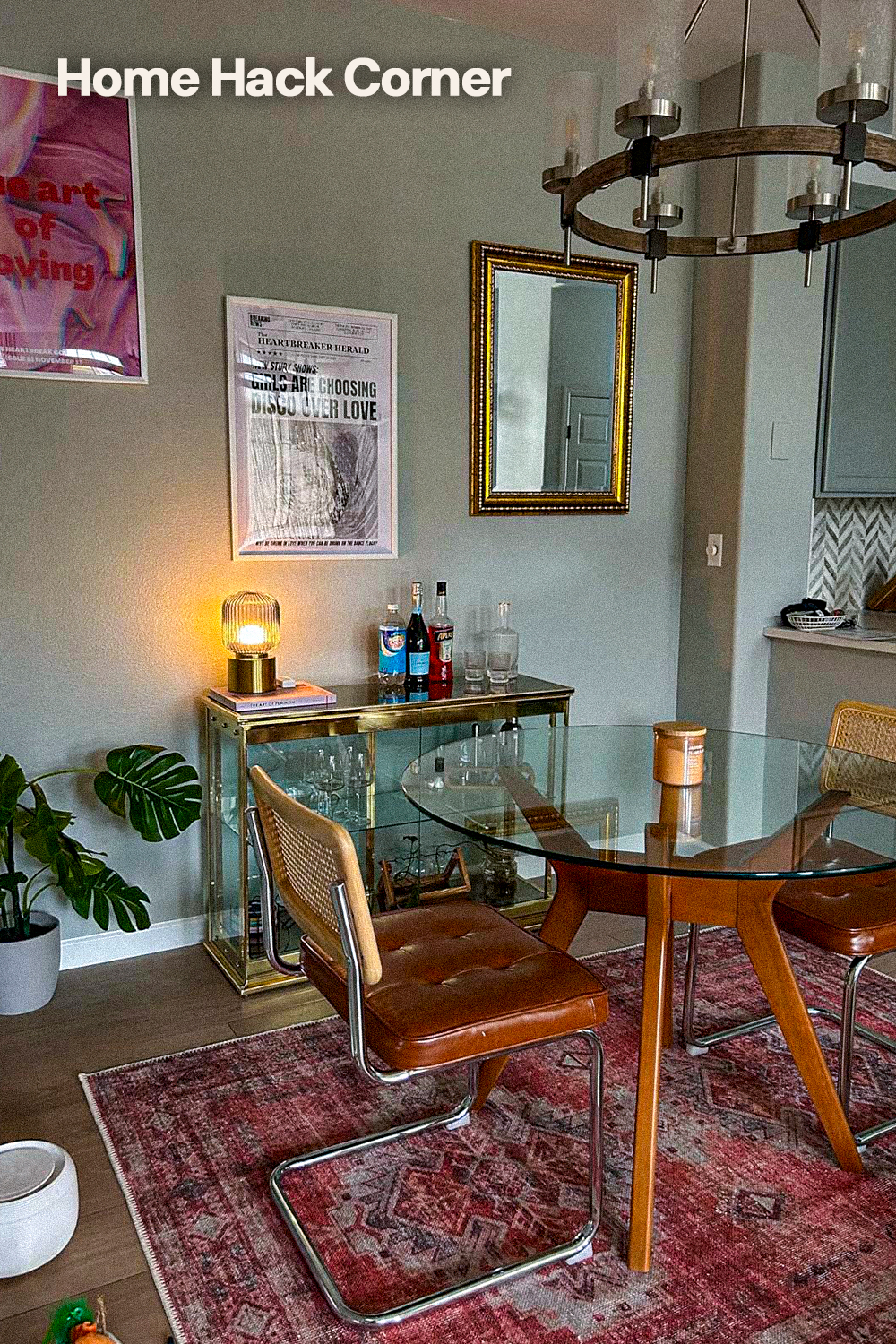Round glass dining table on a splayed wood base with cane back cantilever chairs in tan leather, on a faded red Persian-style rug, beside a brass and glass bar cabinet, under a wagon wheel chandelier