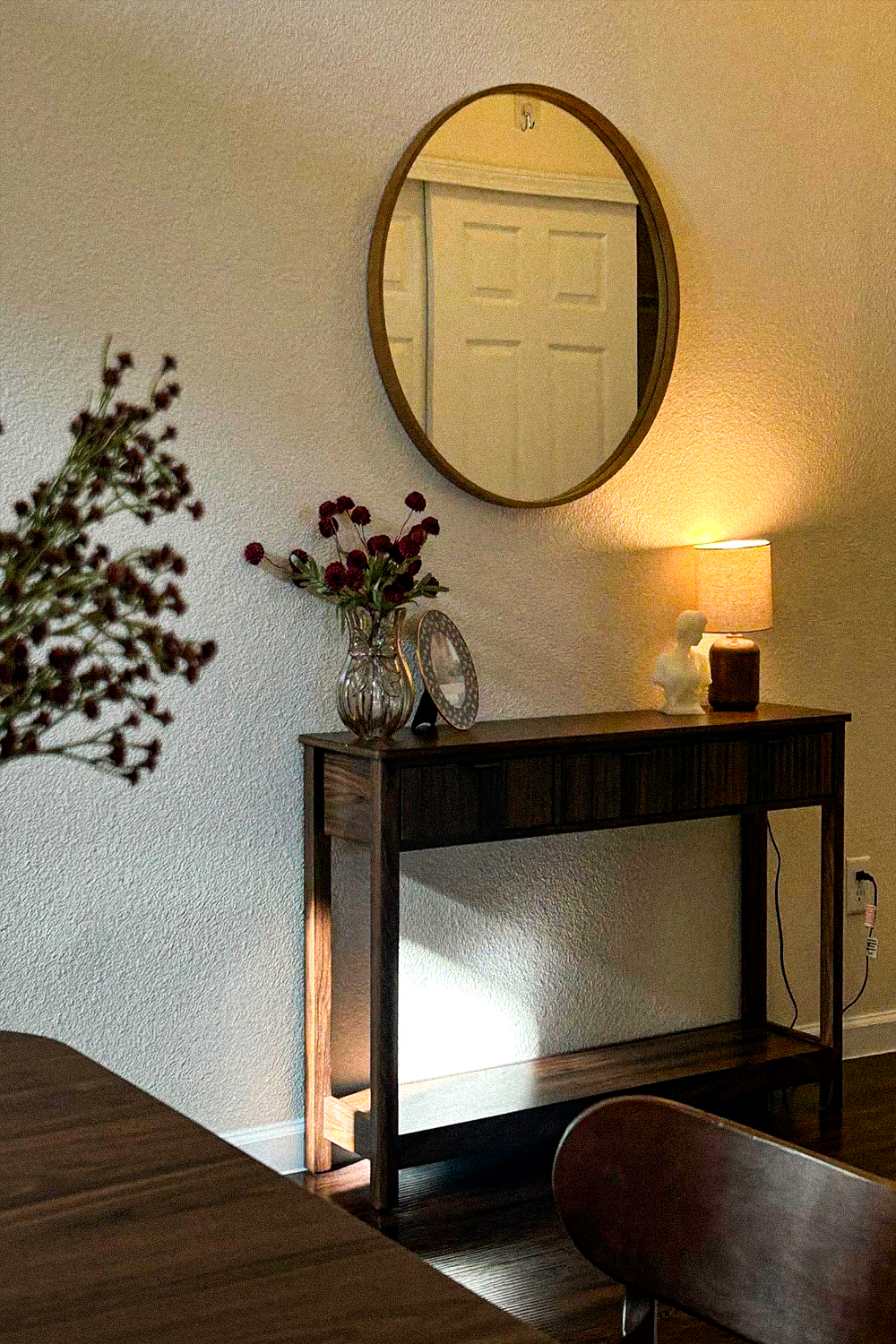 Slim dark wood console table with a fluted drawer against a wall behind a dining table, with a round wood-framed mirror above, a ceramic table lamp, a small marble bust and a glass vase of dried burgundy flowers