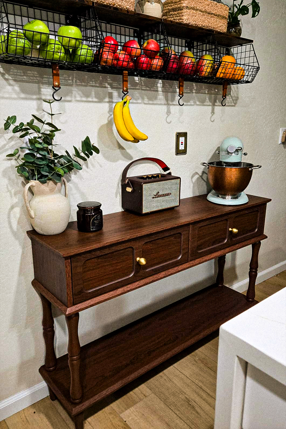 Dark wood console table with turned legs, two drawers with brass knobs and a lower shelf, used as a kitchen sideboard under a wall-mounted shelf of wire fruit baskets with bananas hanging from leather straps, a mint stand mixer with a copper bowl, a retro speaker and a white pitcher of eucalyptus