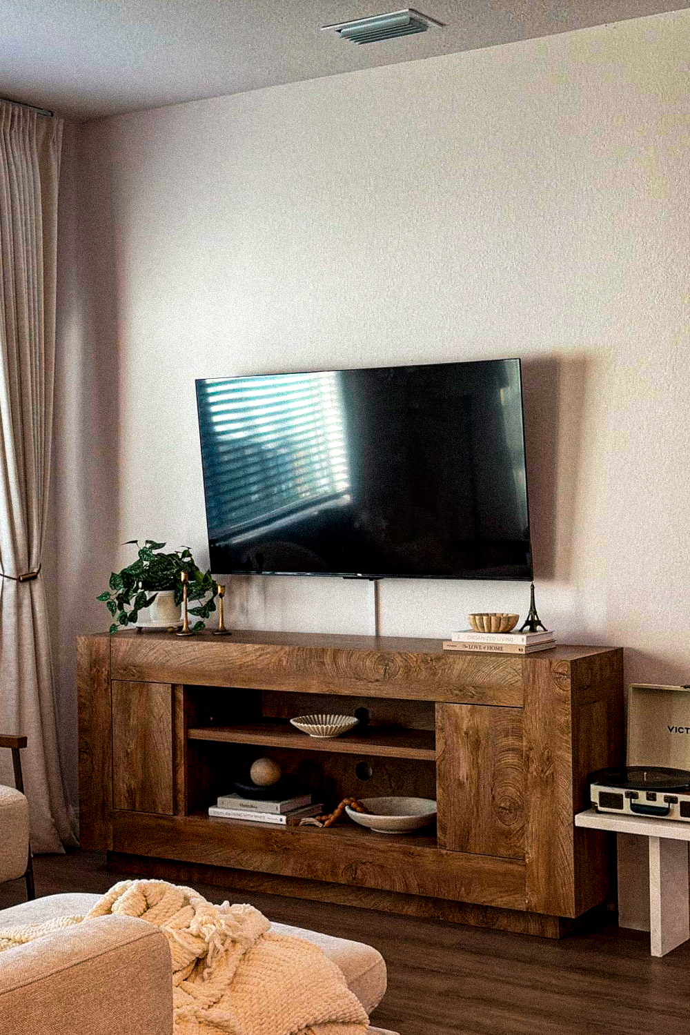 Rustic wood tv stand with cabinet doors on both sides and open shelves in the middle, holding ceramic bowls, a wood bead garland and stacked books, under a wall-mounted tv, with a potted plant on one end, a record player on a small white stand beside it, sheer curtains and a cream knit throw on an ottoman